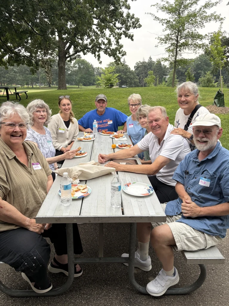 A group of nine smiling adults gathered around a picnic table in a park, enjoying food and conversation outdoors. Trees and open green space surround the group, creating a relaxed and welcoming community setting.