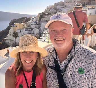 A smiling couple poses for a selfie outdoors on a sunny hillside overlooking white buildings and cliffs along a coastal village. The woman wears a straw hat and pink sleeveless top, while the man wears a patterned shirt and white baseball cap. Other tourists and scenic architecture appear in the background.