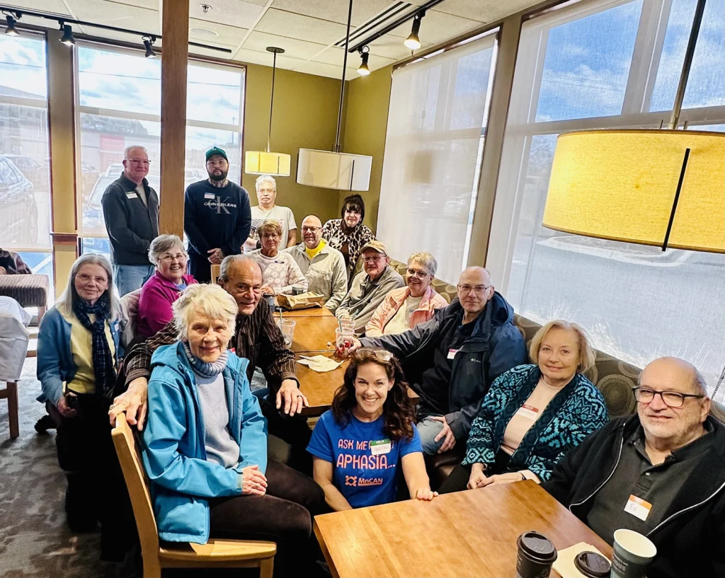 A large group of adults gathers around connected wooden tables inside a café with tall windows and hanging lights. Some people are seated while others stand behind the group, smiling toward the camera. Coffee cups, drinks, and small bags rest on the tables, creating a warm and social community atmosphere.