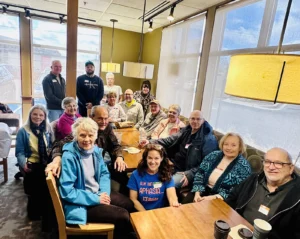 A large group of adults gathers around connected wooden tables inside a café with tall windows and hanging lights. Some people are seated while others stand behind the group, smiling toward the camera. Coffee cups, drinks, and small bags rest on the tables, creating a warm and social community atmosphere.