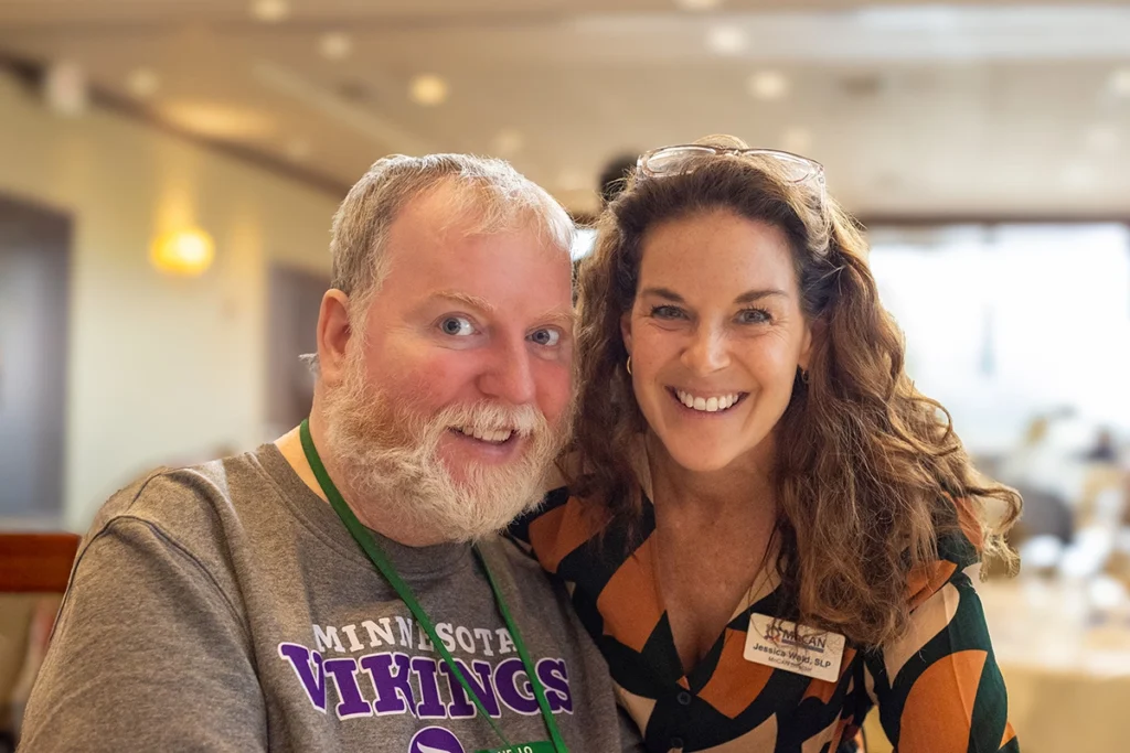 A smiling man with a white beard wearing a Minnesota Vikings T-shirt poses beside a smiling woman with curly hair wearing a patterned blouse and a MnCAN name badge. The two sit closely together indoors in a softly lit setting with a blurred background, creating a warm and friendly atmosphere.