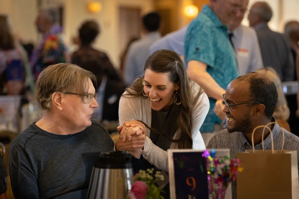 Three people share a warm, joyful moment at a crowded social event. A woman leaning between two seated men smiles while holding one man’s hand as they laugh together. Tables with flowers, gift bags, and coffee carafes fill the foreground, while other guests mingle in the softly blurred background.