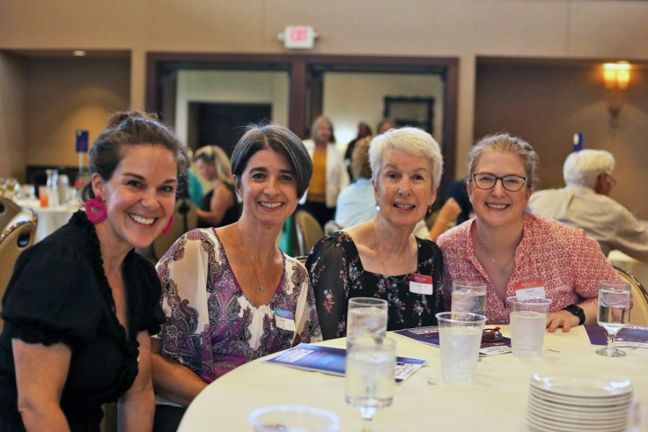 Four women sit together at a round banquet table smiling toward the camera during a community or social event. Glasses of water, plates, and printed programs rest on the table, while other attendees and softly lit meeting-room décor appear in the background.