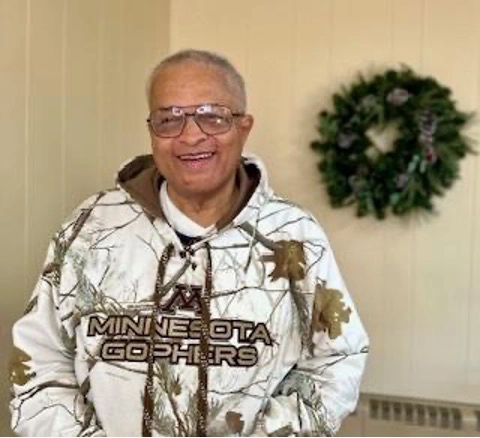 A smiling man wearing glasses and a white Minnesota Gophers camouflage hoodie stands indoors in front of a light-colored wall with a holiday wreath hanging in the background.