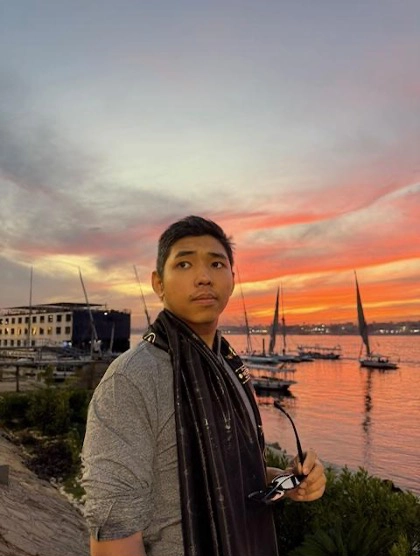 A young man stands near a waterfront at sunset, holding sunglasses and looking off into the distance. Sailboats float on calm water behind him, while the sky glows with orange, pink, and purple sunset colors.