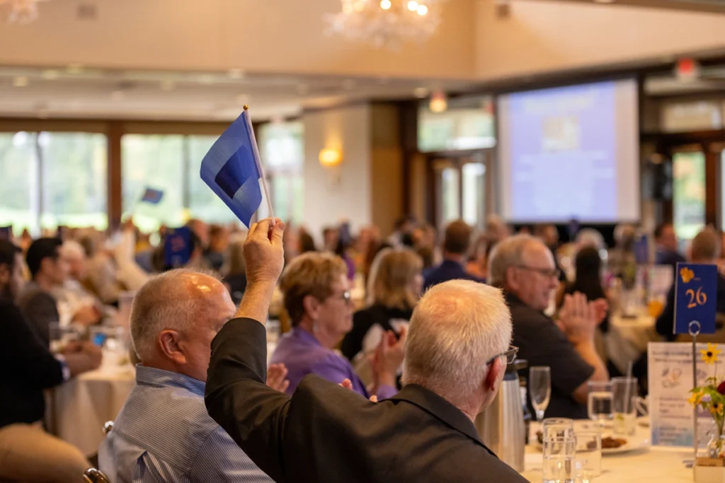 A large audience gathered at a banquet-style event, with one attendee raising a blue card or flag while others applaud. Round tables fill the room, and a presentation screen is visible in the background. The atmosphere feels engaged and celebratory.