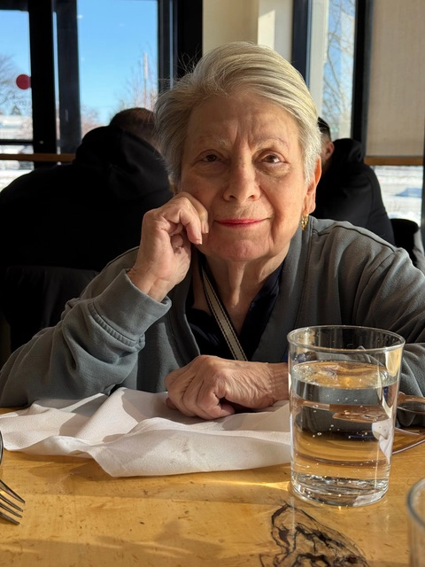 An older woman with short gray hair sits at a restaurant table, resting her chin on one hand and smiling gently toward the camera. A glass of water and folded napkin sit on the table in front of her, while sunlight streams through large windows in the background.